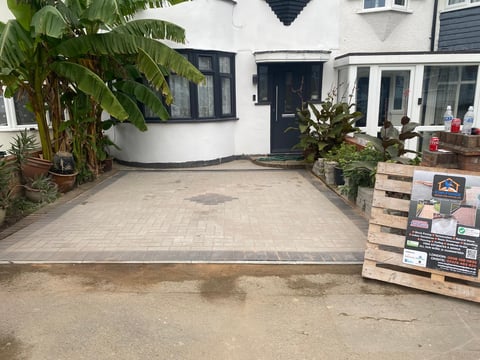 A newly laid paved driveway in front of a modern white house with green plants and wooden pallets stacked on the right side