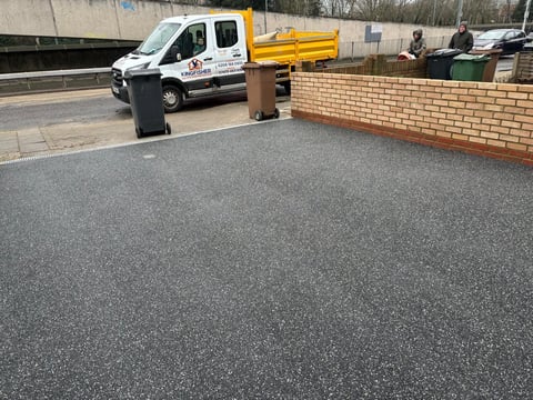 Newly paved asphalt driveway with white and yellow work truck and brick wall in background