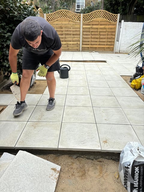Man in black shirt installing paving stones in a garden patio, with wooden lattice fence in background