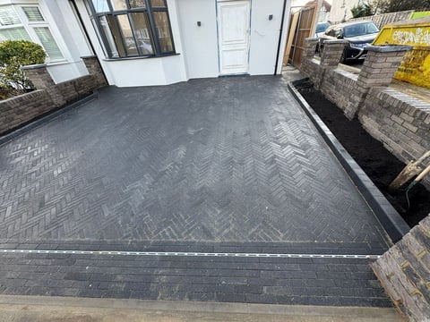 New dark herringbone pattern driveway with white door and house entrance, bordered by stone walls